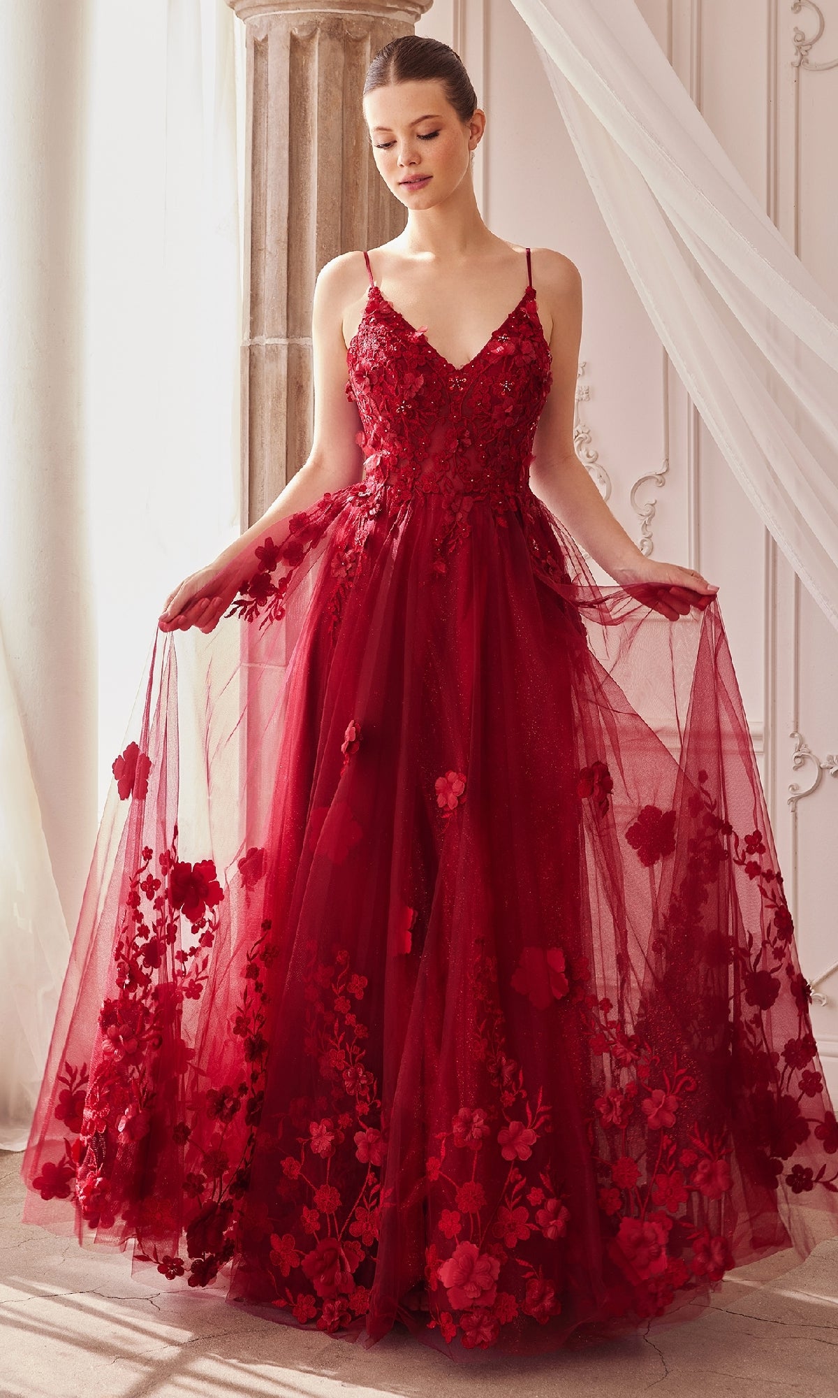 A woman stands indoors, gracefully holding out the sides of her Long Glitter Prom Ball Gown: Andrea and Leo A1326—a sleeveless red formal dress with floral appliqués and a tulle skirt.